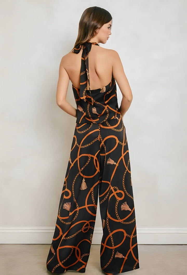 A woman modeling a black wide-leg halter jumpsuit from the back, featuring an orange chain-link and rope pattern, set against a plain light gray wall.