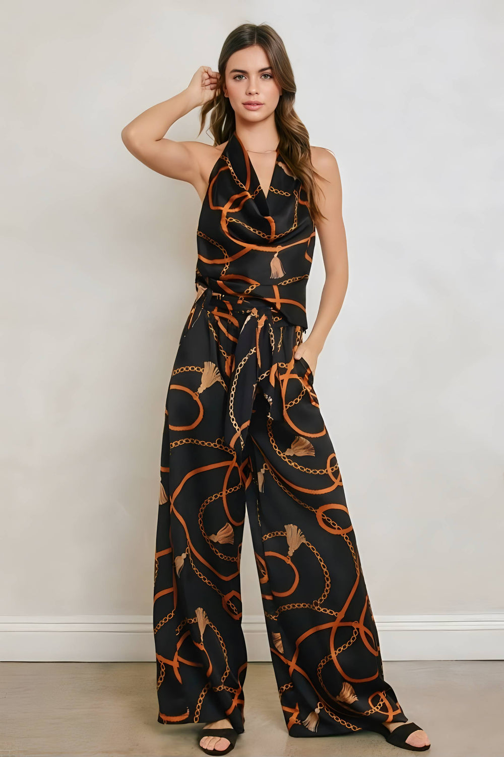 A woman modeling a sleeveless halter-neck jumpsuit with a black base and an orange chain-link pattern, standing against a light gray wall.