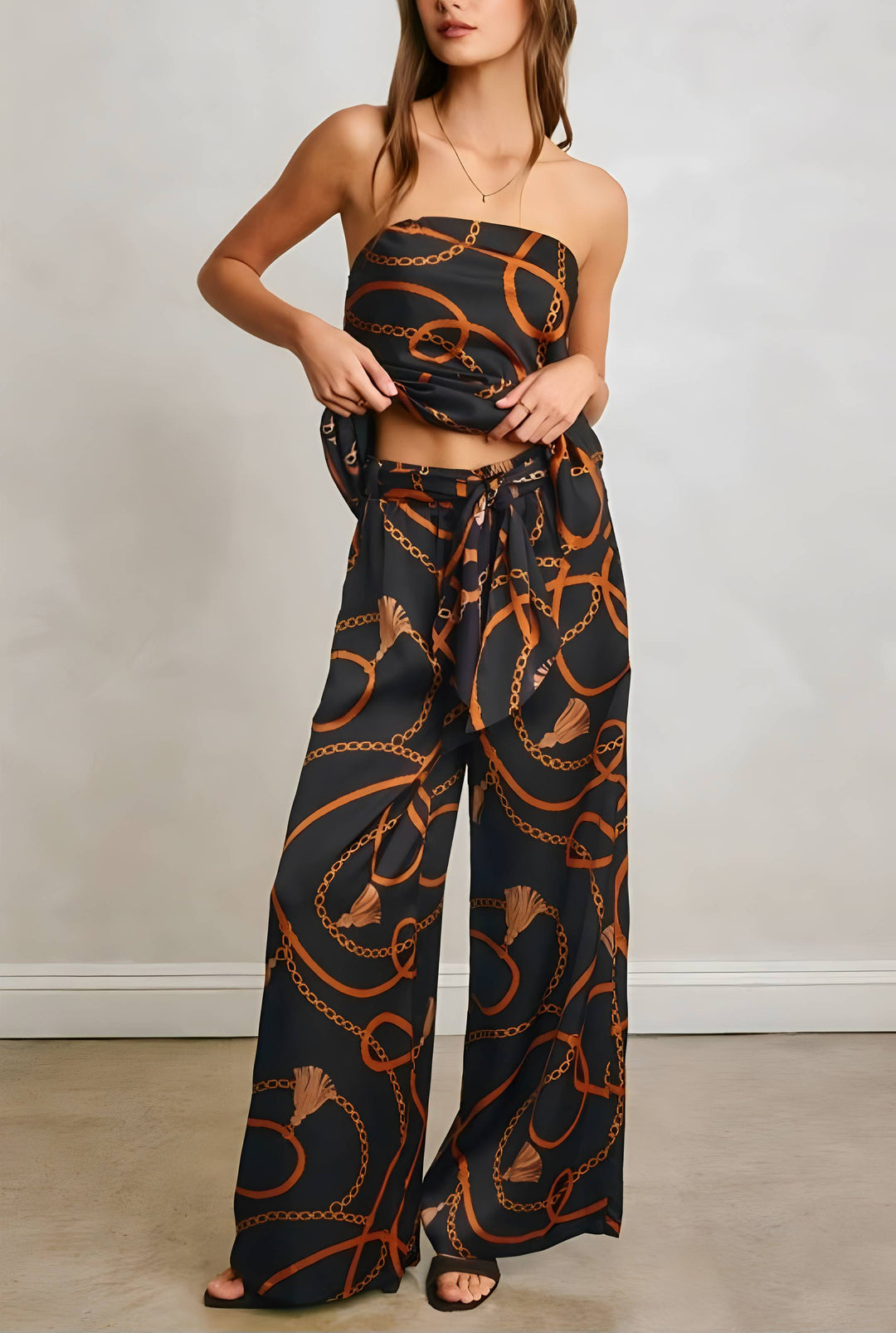 A woman models a matching strapless bandeau top and wide-leg pants set in black with an orange chain-link pattern, standing against a neutral light gray wall.