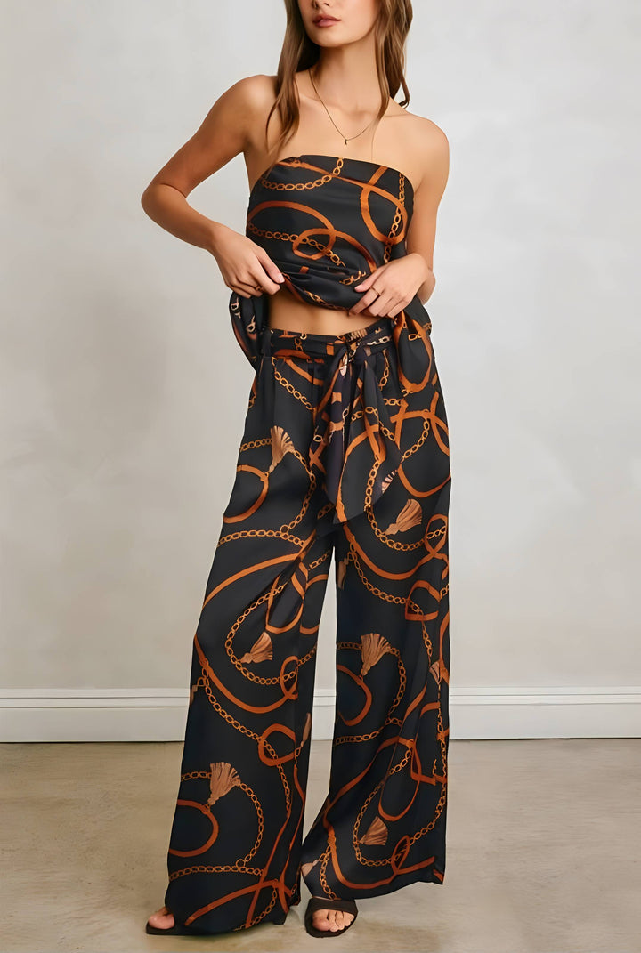 A woman models a matching strapless bandeau top and wide-leg pants set in black with an orange chain-link pattern, standing against a neutral light gray wall.