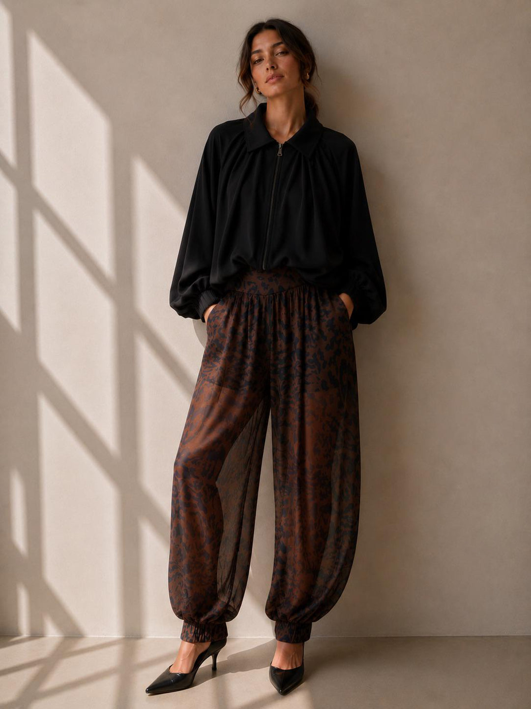 A woman stands against a beige wall wearing a black long-sleeve collared top and loose brown patterned harem-style pants with black pointed heels. Sunlight casts window-frame shadows on the wall.