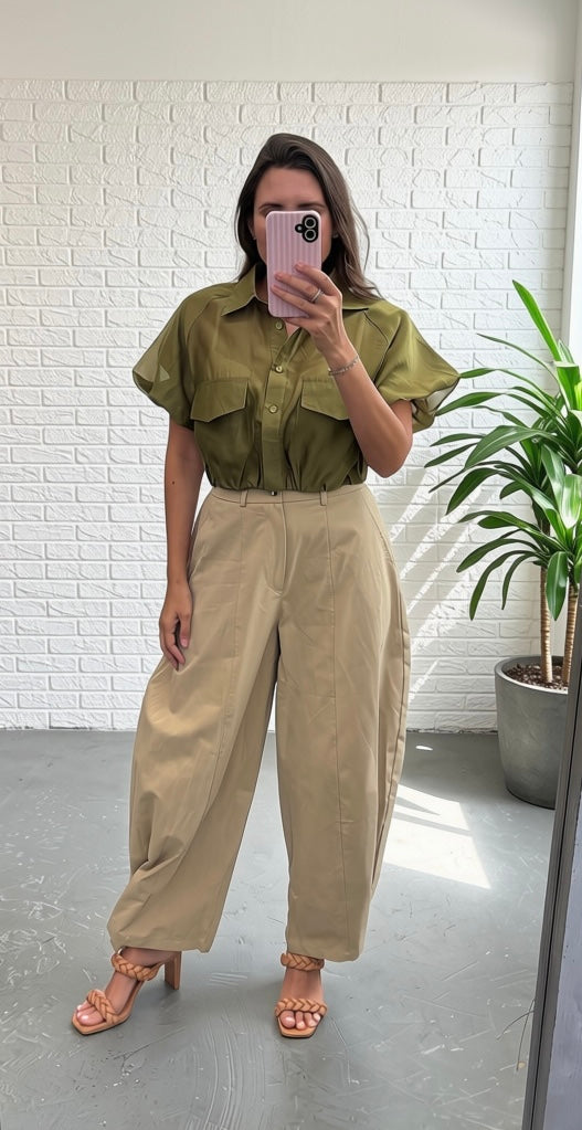 A woman takes a mirror selfie wearing an olive green short-sleeve button-up shirt tucked into high-waisted, wide-leg beige trousers, paired with tan heeled sandals. She is standing in a room with a white brick wall and a potted plant.