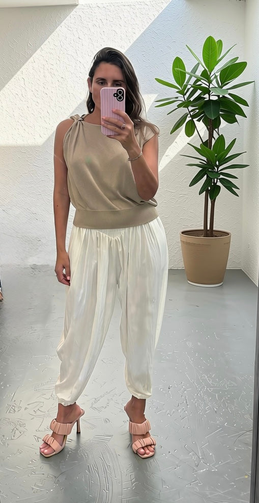 A woman takes a mirror selfie wearing a beige halter top and white satin harem pants with pink heeled sandals, standing in a bright room with a potted plant.