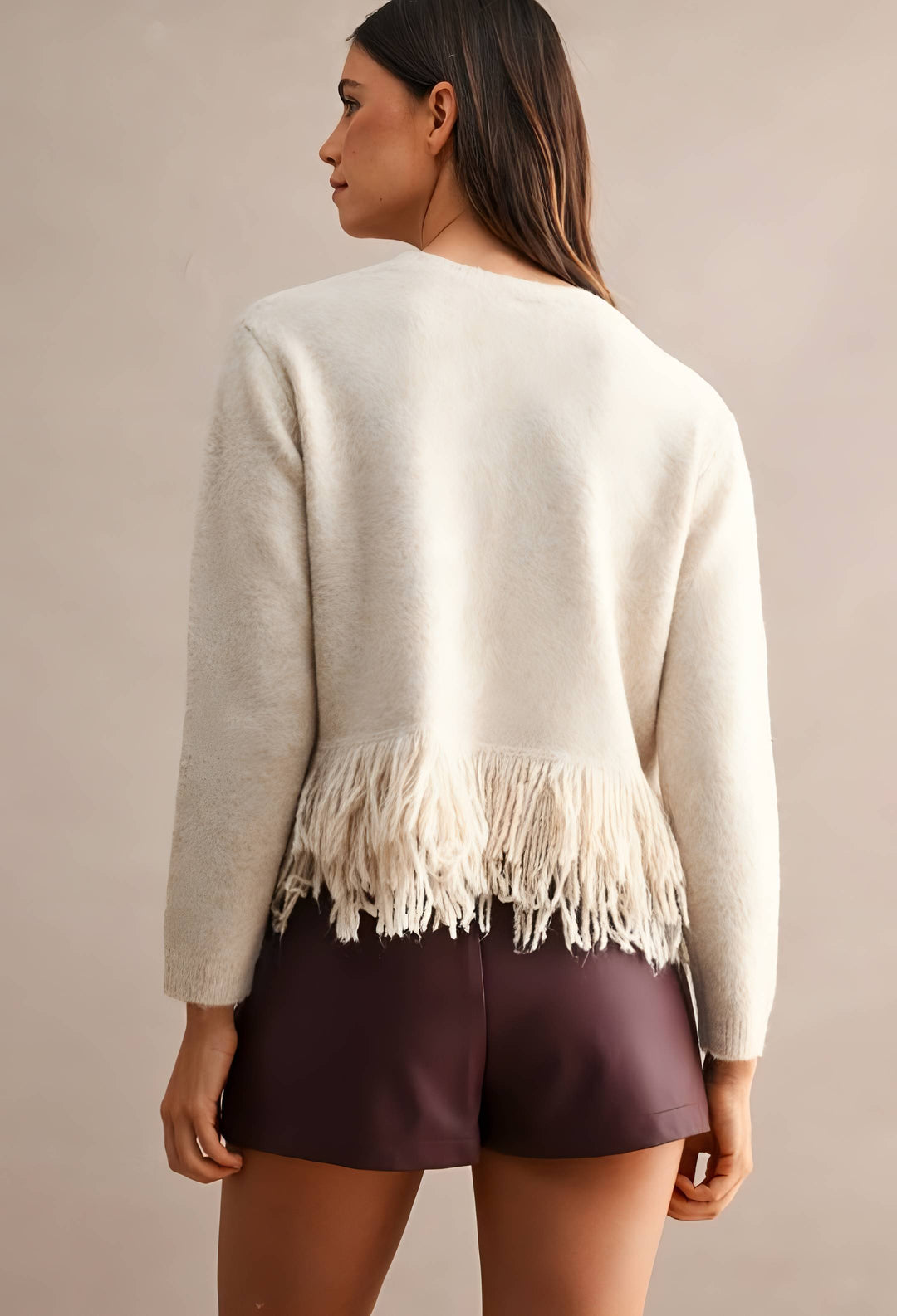 A woman shown from the back wearing a cream-colored knit sweater with fringe trim at the hem and dark plum shorts, set against a plain beige studio background.