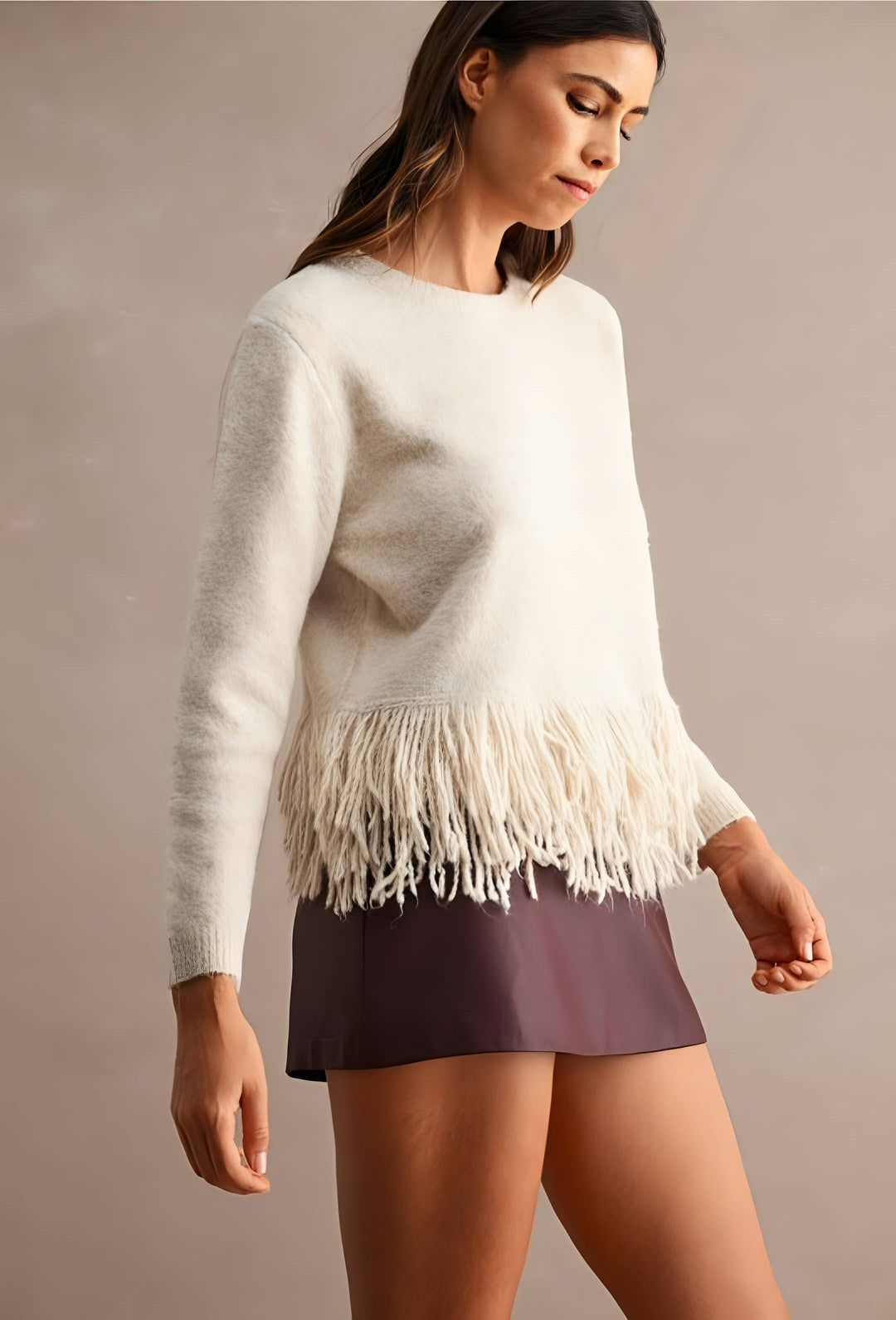 A woman wearing a cream-colored knit sweater with a fringe hem and dark plum-colored shorts, standing against a neutral beige studio background.
