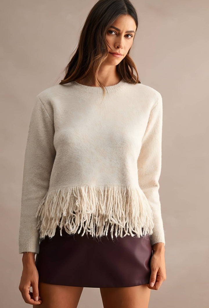 A woman wearing a cream-colored knit sweater with a fringe hem, paired with a dark purple mini skirt, against a solid beige studio background.
