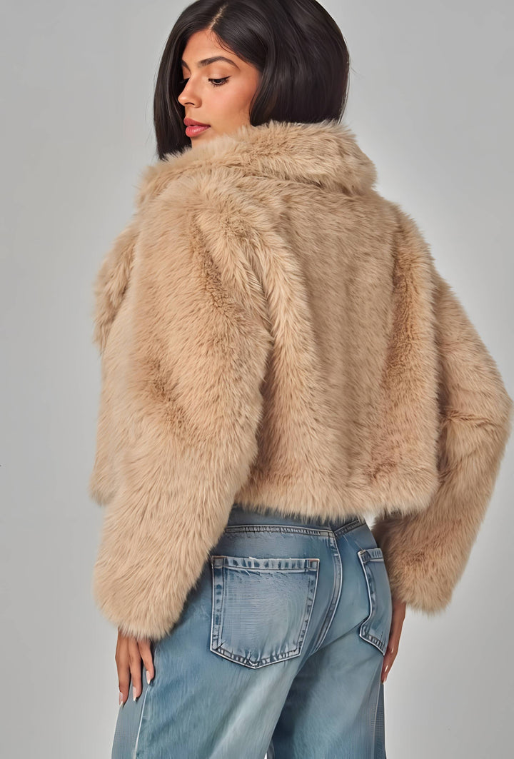 A woman shown from the back wearing a cropped, plush faux-fur jacket in a light beige/tan shade, paired with light-wash blue jeans, against a neutral gray studio background.