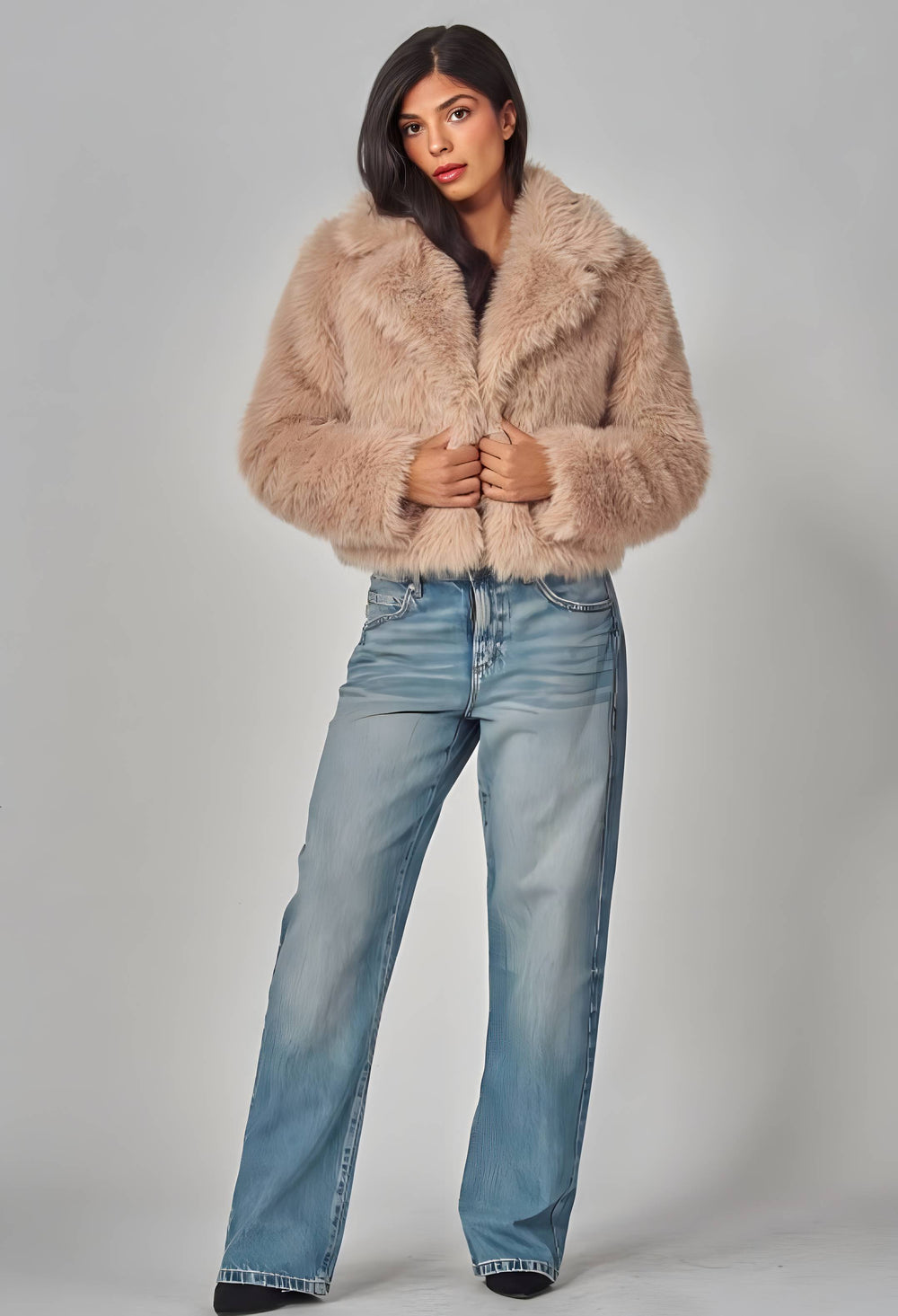 A woman modeling a cropped, light beige faux fur jacket with a wide collar, paired with light-wash blue jeans, against a neutral gray studio background.