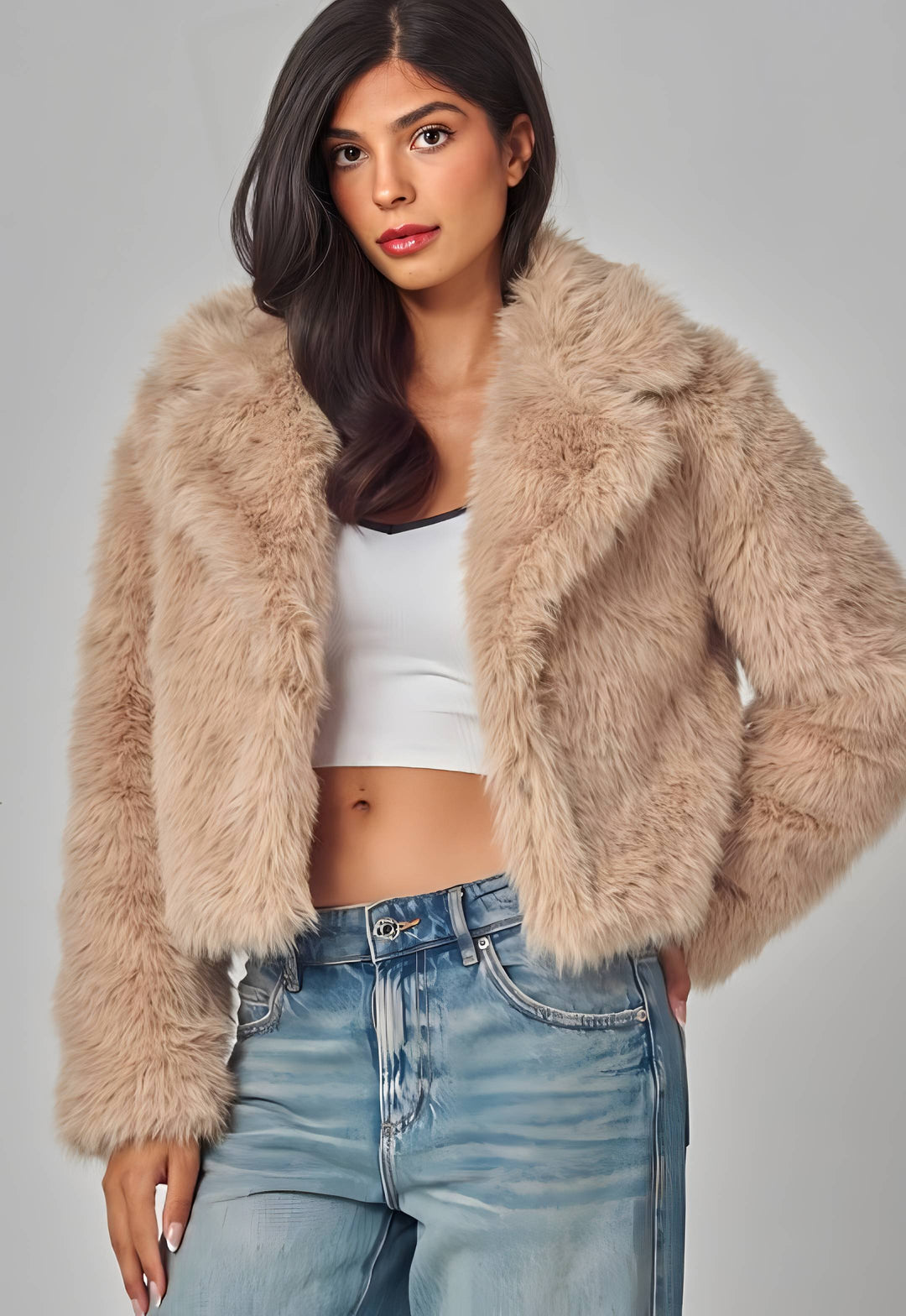 A woman modeling a cropped, plush faux fur jacket in a light beige or taupe color, worn over a white crop top and light-wash blue jeans against a neutral gray studio background.