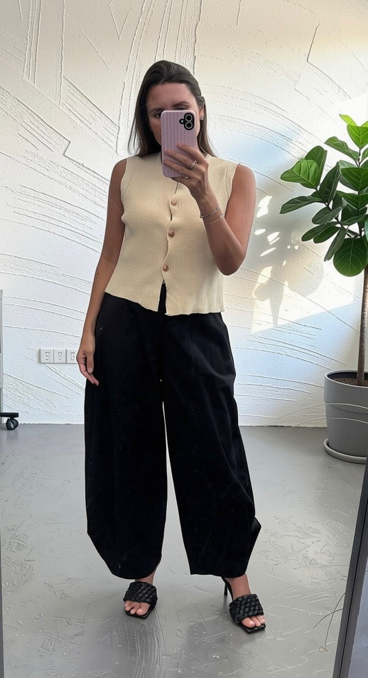 A woman takes a mirror selfie wearing a sleeveless cream-colored button-up vest and black wide-leg pants, paired with black woven slide sandals, in a bright room with a potted plant.