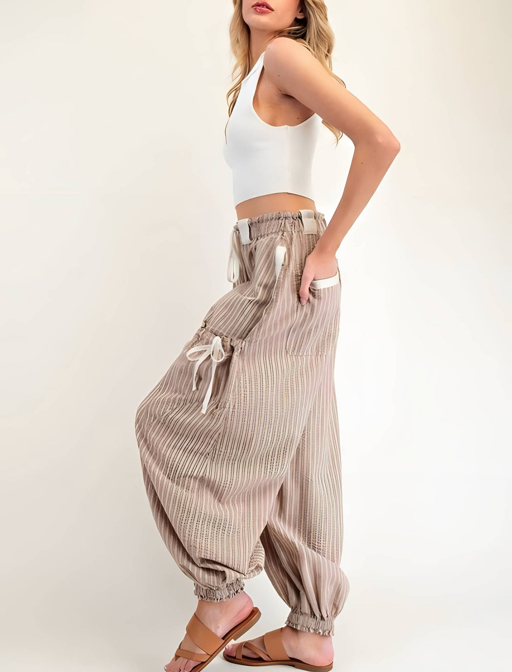 A woman modeling loose, high-waisted striped harem pants in beige and white, paired with a white crop top and brown flat sandals, against a plain off-white studio background.