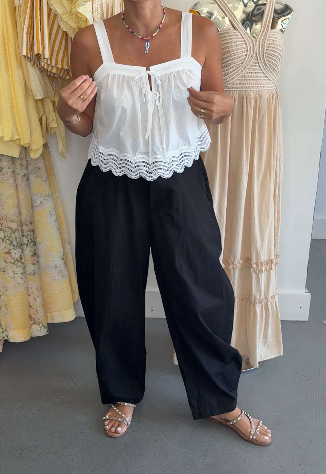 A woman modeling a white sleeveless top with a scalloped wavy hem and black wide-leg pants, standing in a clothing store next to mannequins wearing yellow and beige dresses.