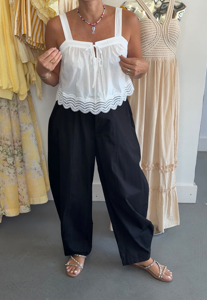 A woman modeling a white sleeveless top with a scalloped wavy hem and black wide-leg pants, standing in a clothing store next to mannequins wearing yellow and beige dresses.