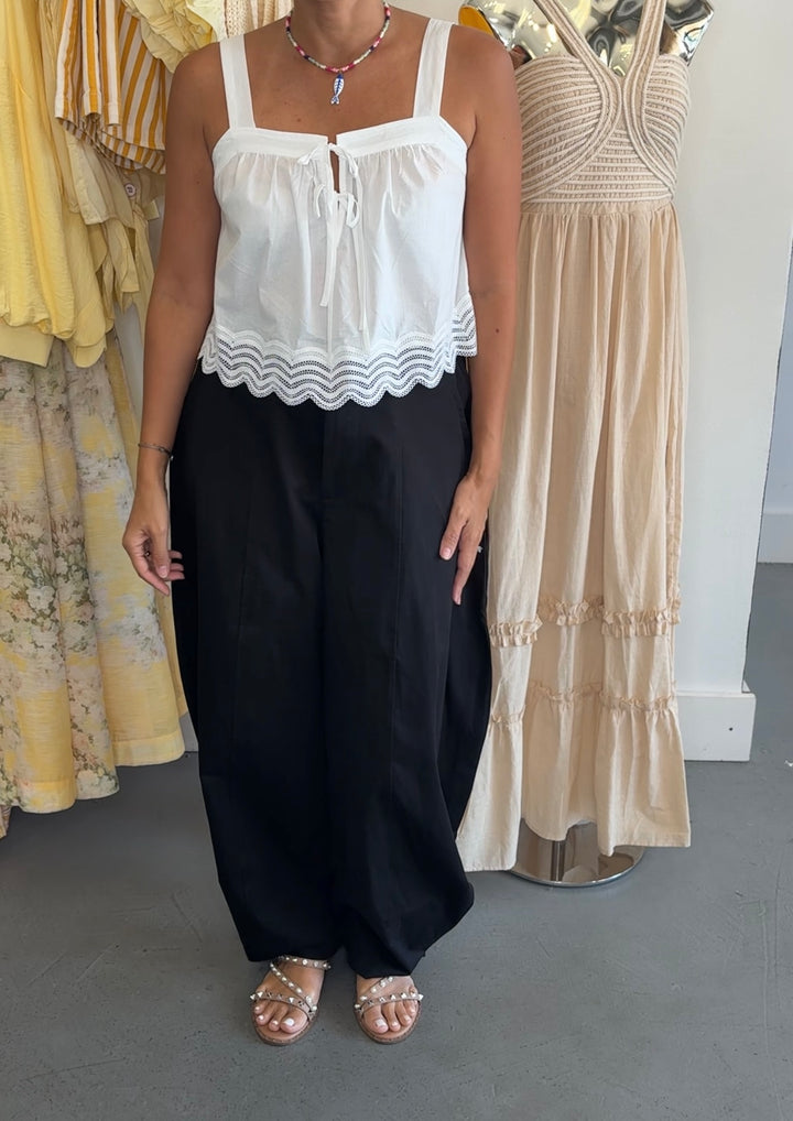 A woman modeling a white sleeveless top with a scalloped hem and black wide-leg pants, standing in a clothing store next to mannequins wearing beige and yellow dresses.