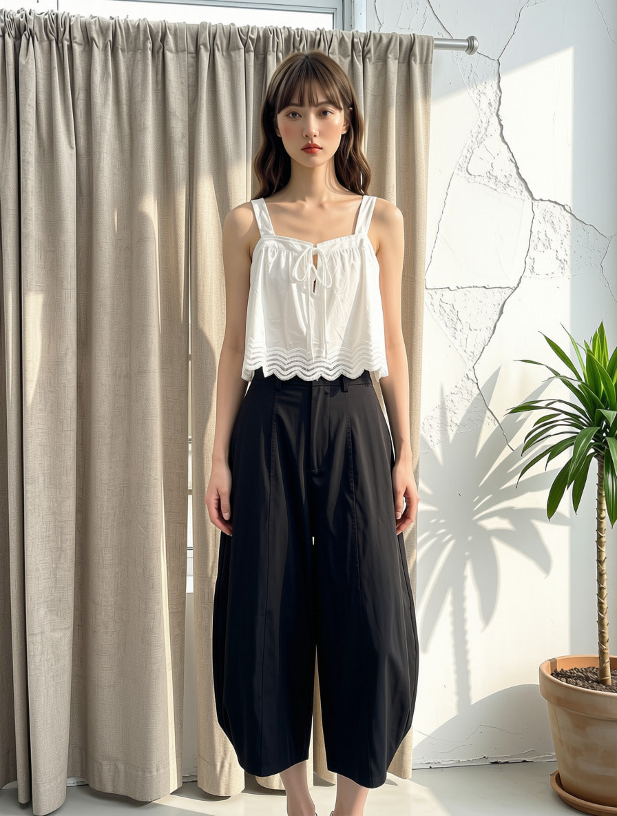 A woman stands indoors wearing a white sleeveless top with a ruffled hem and black wide-leg cropped pants. She is positioned in front of beige curtains, a white wall with wall cracks, and a potted plant.