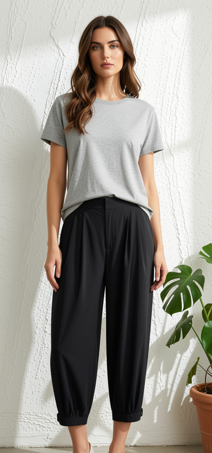 A woman modeling a light gray short-sleeve crewneck t-shirt and black pleated cropped pants, standing against a textured white wall with a potted plant on the right.