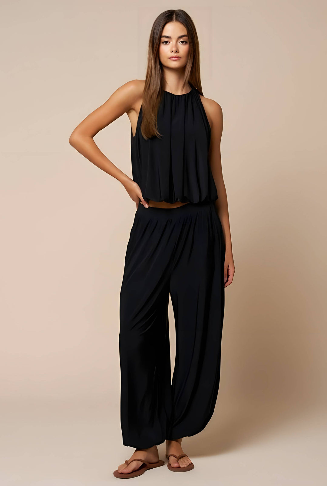 A woman modeling a black sleeveless halter top and matching wide-leg pants set, paired with brown sandals, against a solid beige studio background.