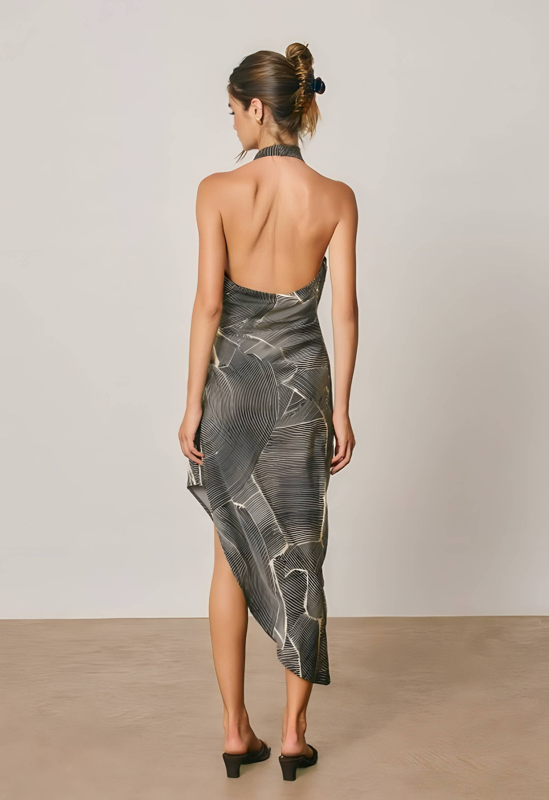 A woman stands with her back to the camera wearing a sleeveless, open-back midi dress with an abstract gray and black pattern and a halter neckline. She is photographed in a minimalist studio setting with a light gray wall and beige floor.