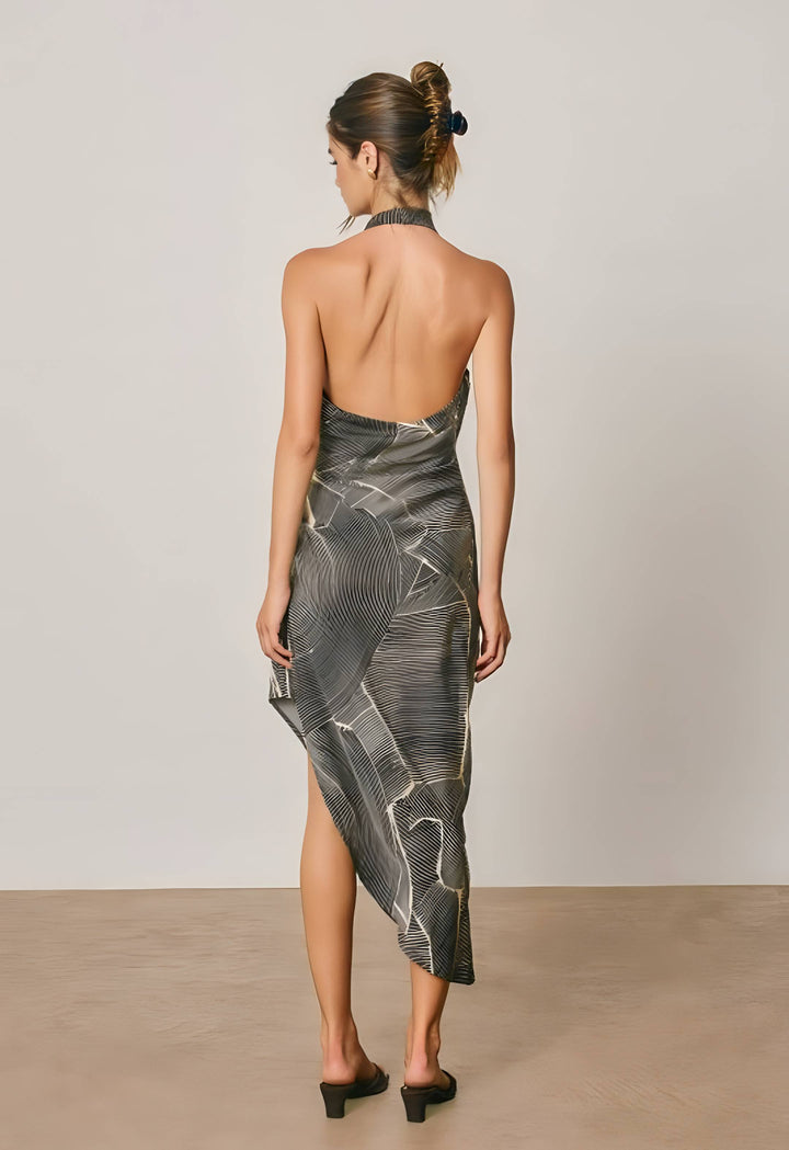 A woman stands with her back to the camera wearing a sleeveless, open-back midi dress with an abstract gray and black pattern and a halter neckline. She is photographed in a minimalist studio setting with a light gray wall and beige floor.
