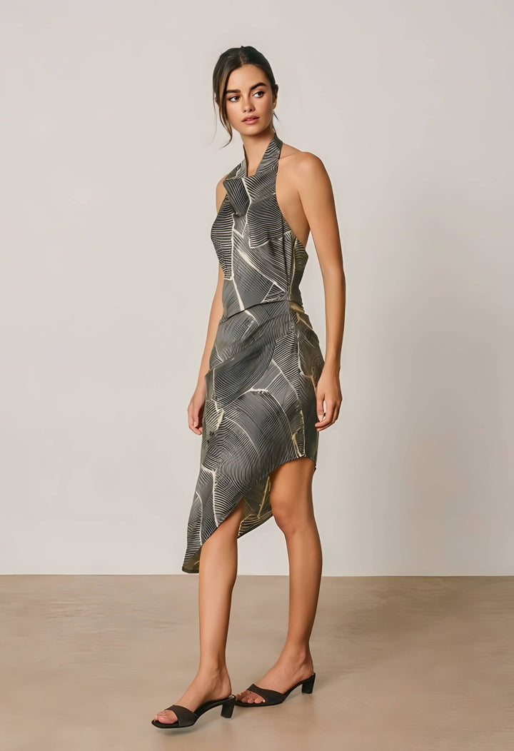 A woman modeling a sleeveless, high-neck wrap-style midi dress with a gray, black, and white abstract line pattern, paired with black slide heels, set against a neutral studio backdrop.