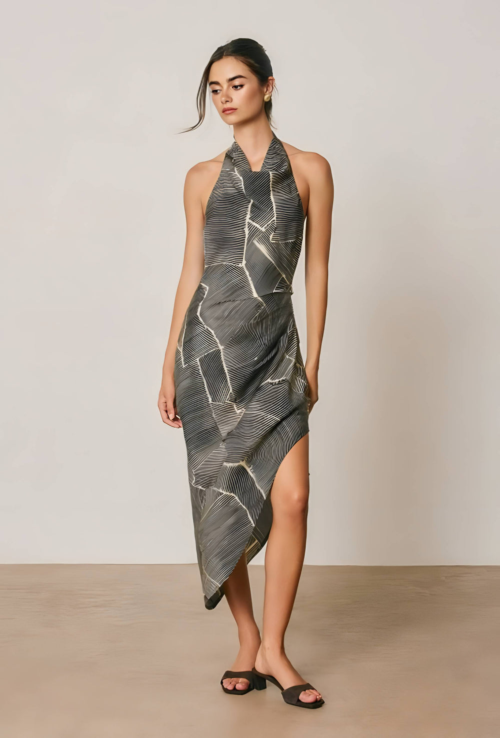 A model wearing a sleeveless halter-neck midi dress with a gray and black abstract line pattern, standing against a neutral beige studio background.