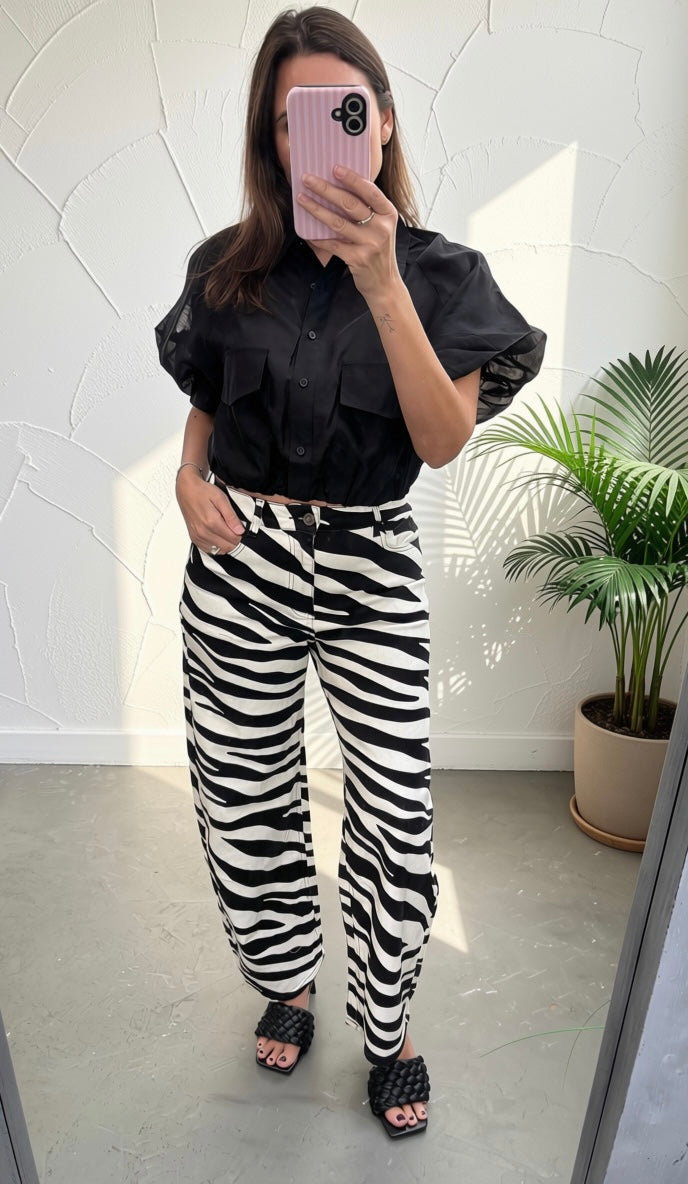 A woman takes a mirror selfie wearing a black short-sleeve button-up shirt and black-and-white zebra print wide-leg pants, paired with black woven slide sandals, in a bright room with a potted plant.
