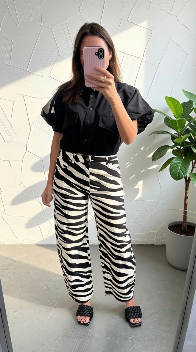 A woman takes a mirror selfie wearing a black puff-sleeve button-up shirt and high-waisted black and white zebra-print wide-leg trousers, paired with black slide sandals, against a textured white wall with a potted plant.