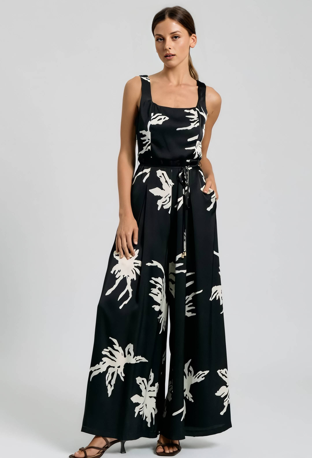 A woman modeling a sleeveless black wide-leg jumpsuit with a white tropical floral print, photographed against a neutral light gray studio background.
