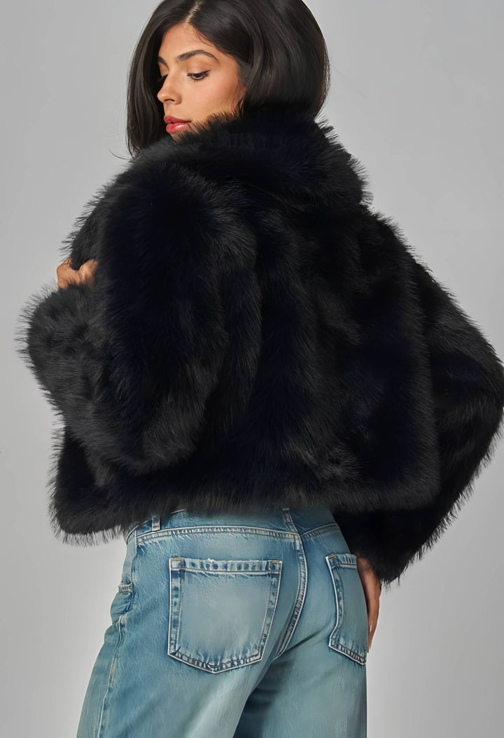 A woman wearing a voluminous black faux fur jacket with wide draped sleeves, paired with light blue jeans, photographed against a neutral gray studio background.