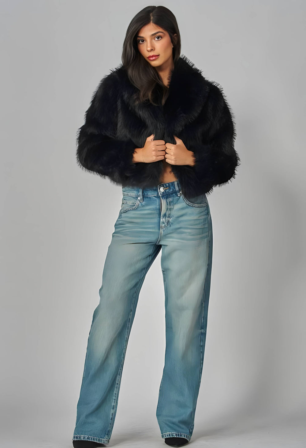 A model wearing a black faux fur cropped jacket and light blue wide-leg jeans, standing against a neutral gray studio background.