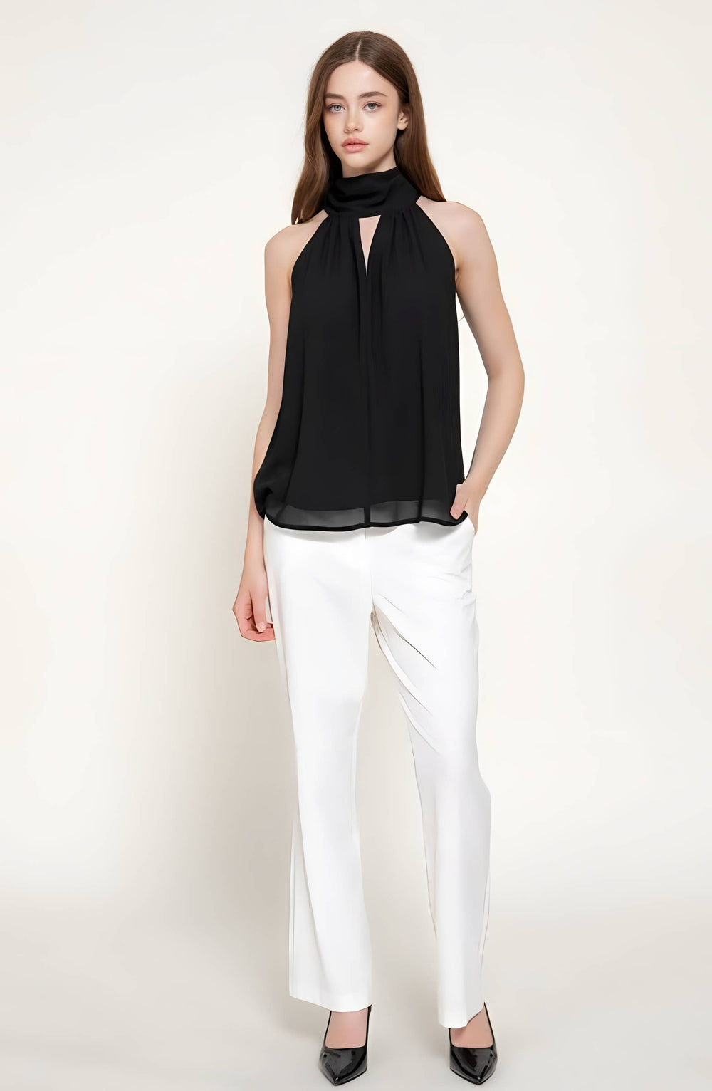 A woman modeling a sleeveless black halter-neck chiffon top paired with white trousers and black pointed shoes, standing against a plain off-white studio background.