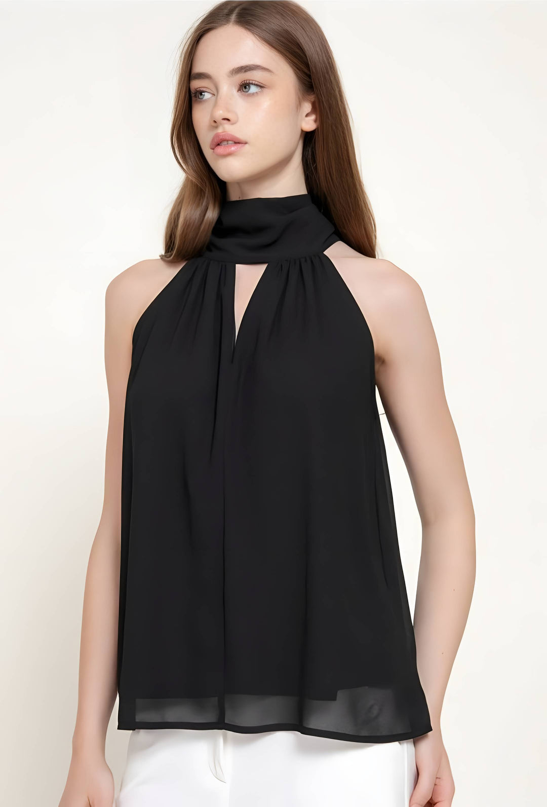 A woman wearing a sleeveless black high-neck blouse with a keyhole cutout and flowing chiffon-like fabric, paired with white pants, against a plain light background.