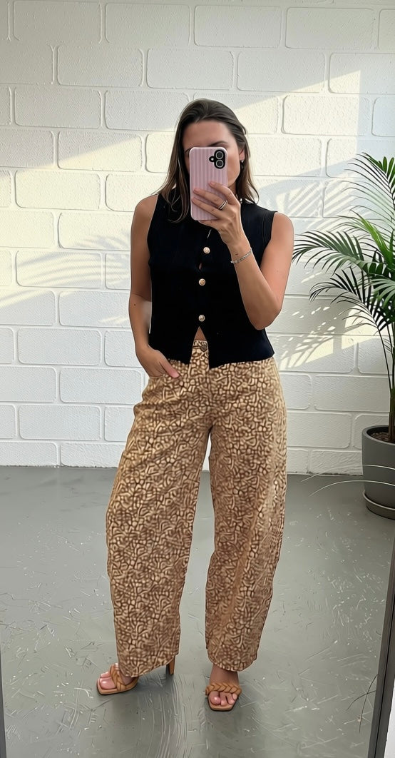 A woman takes a mirror selfie wearing a sleeveless black vest with gold buttons and wide-leg beige pants with a small gold pattern, standing in front of a white brick wall.