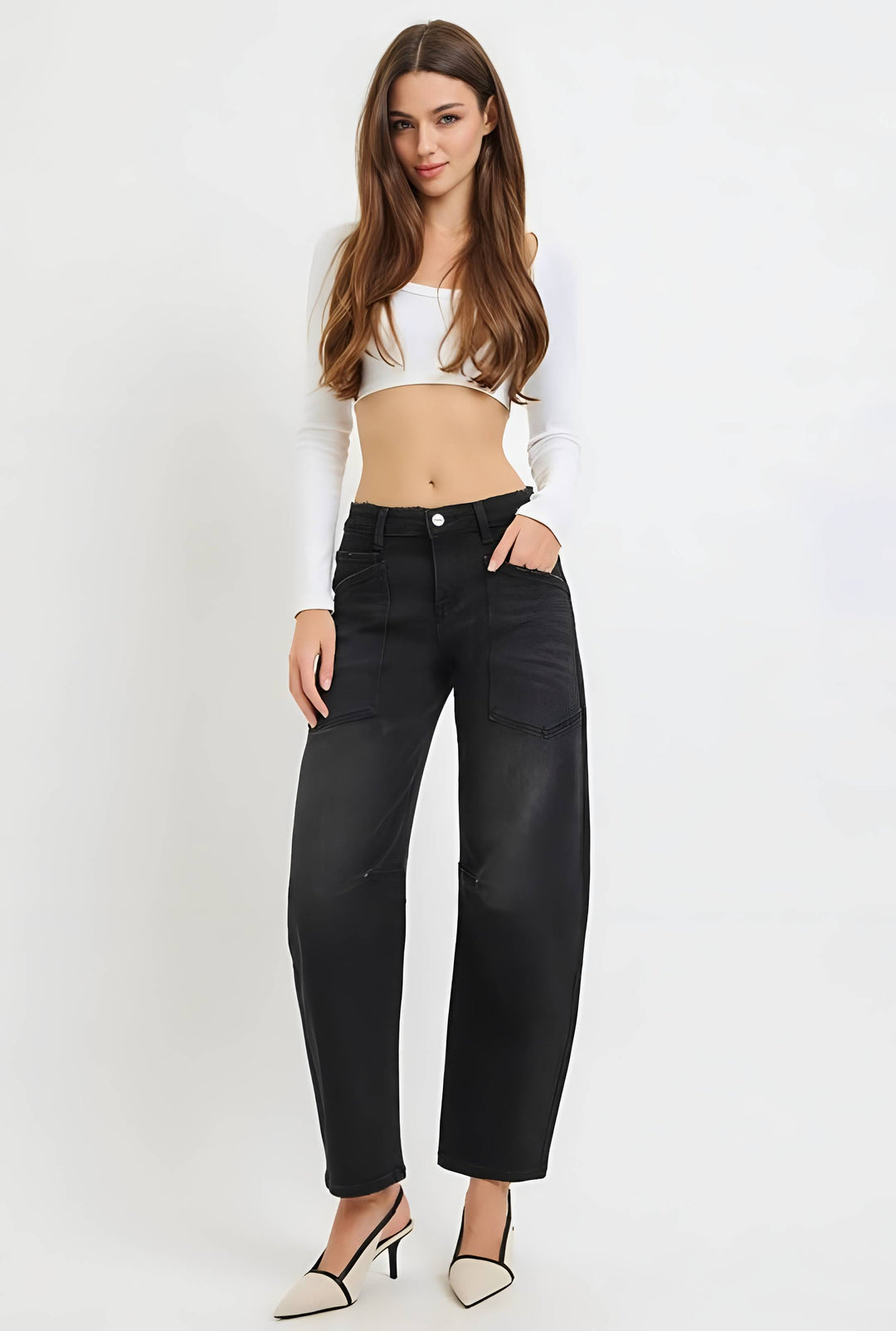 A woman modeling black wide-leg cropped jeans with a white long-sleeve crop top and white pointed-toe heels, standing against a plain white studio background.