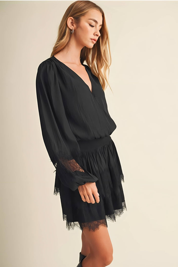 A woman wearing a black long-sleeve mini dress with a deep V-neckline, sheer lace cuffs, and a ruffled tulle hem, posed against a solid light beige background.