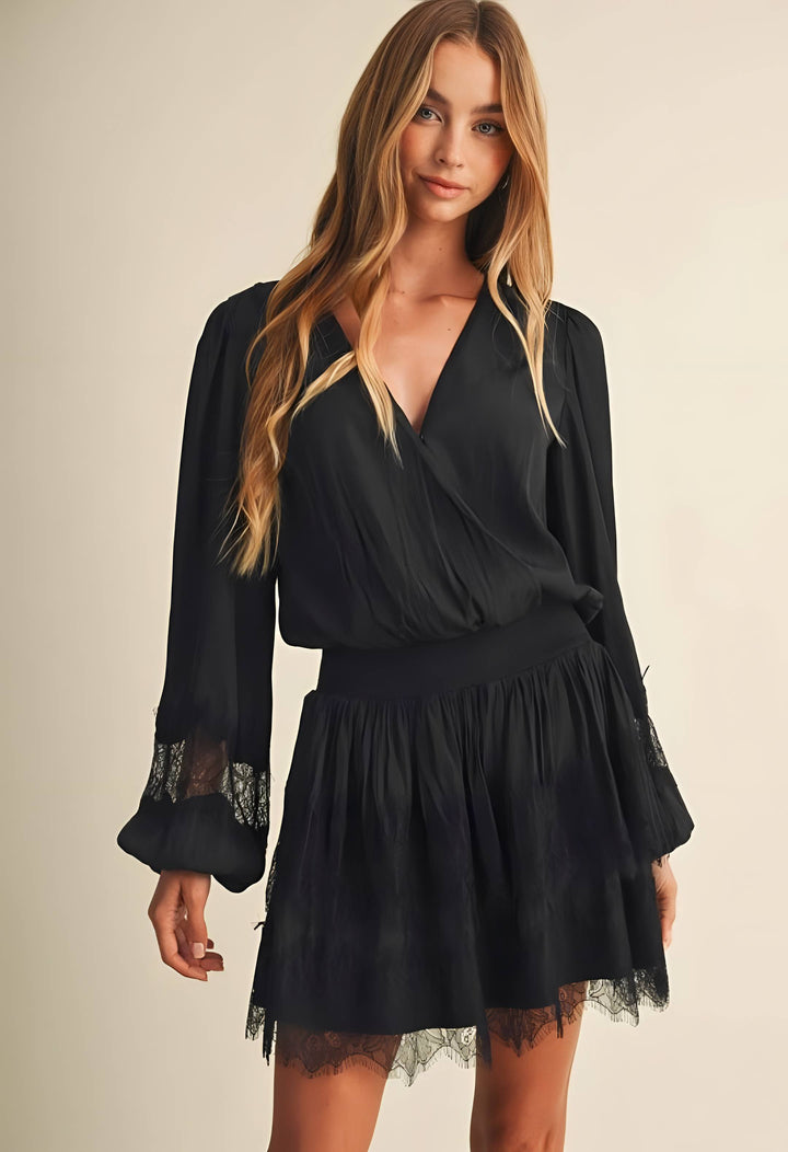 A woman modeling a black long-sleeve wrap-style mini dress with a flared skirt and lace trim on the cuffs and hem, photographed against a plain light beige studio background.