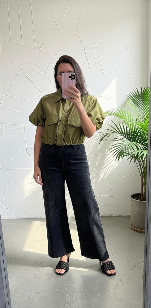 A woman takes a mirror selfie showing an outfit consisting of an olive green short-sleeve button-up shirt and black wide-leg jeans, paired with black woven slide sandals. The background is a modern white textured wall with a potted palm plant on the right.