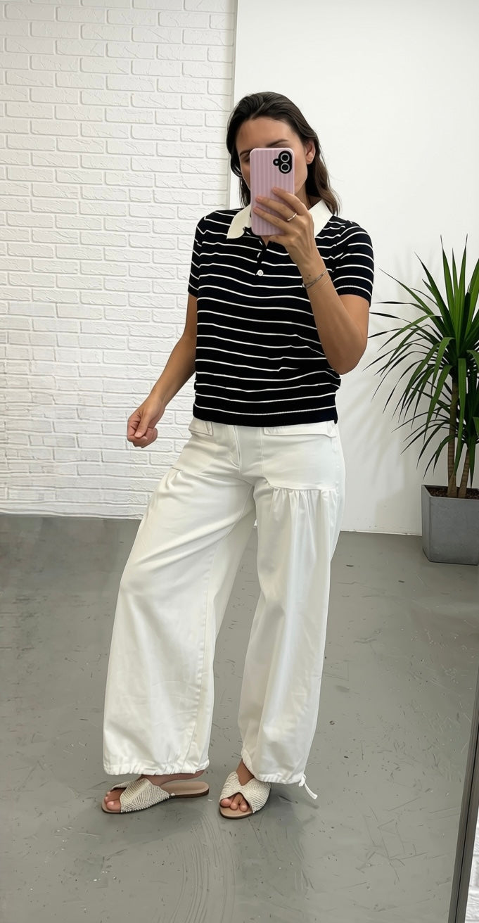 A woman takes a mirror selfie wearing a black and white striped short-sleeve polo shirt with a white collar, paired with wide-leg white pants and beige slide sandals, set against a white brick wall and gray floor.
