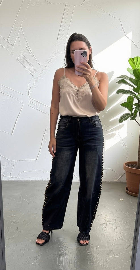 A woman takes a mirror selfie showing off a pair of wide-leg black jeans with silver studs down the sides, paired with a beige camisole top and black sandals.
