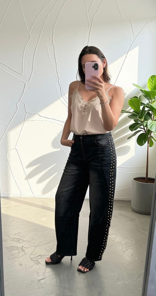 A woman takes a mirror selfie wearing a light beige lace-trimmed camisole top and black wide-leg jeans with silver studs down the side, paired with black heeled sandals.