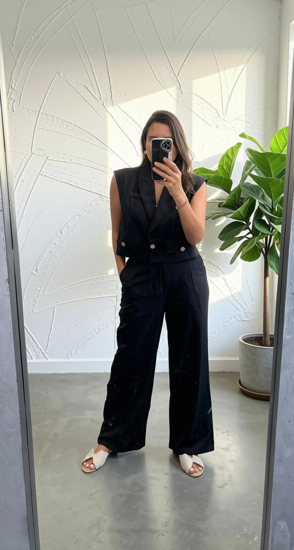 A woman takes a mirror selfie wearing a black sleeveless double-breasted vest and matching wide-leg trousers, paired with white slide sandals. The setting is a modern room with a white textured wall, gray concrete floor, and a green potted plant.