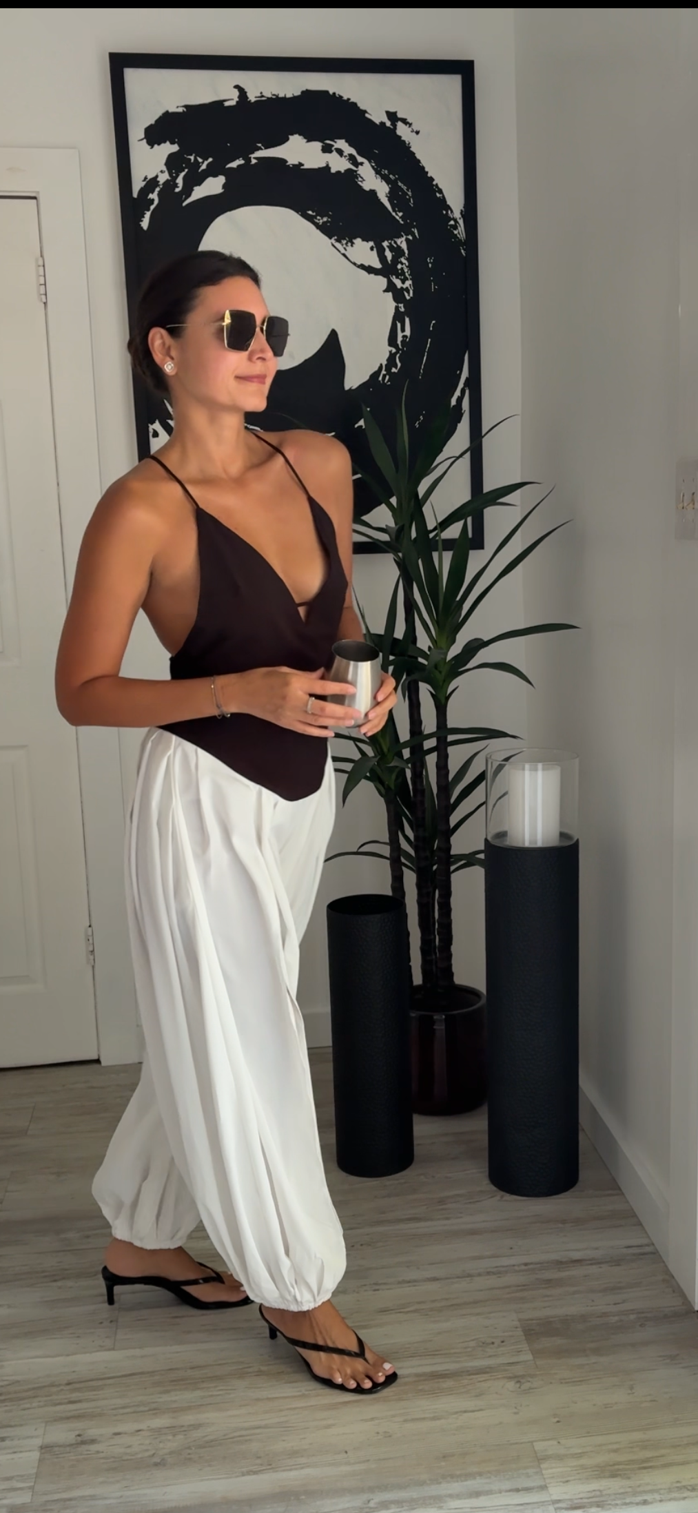 A woman wearing a dark brown halter top and white wide-leg pants stands in a modern indoor setting with a framed black and white wall art piece, a potted plant, and black cylindrical side tables.