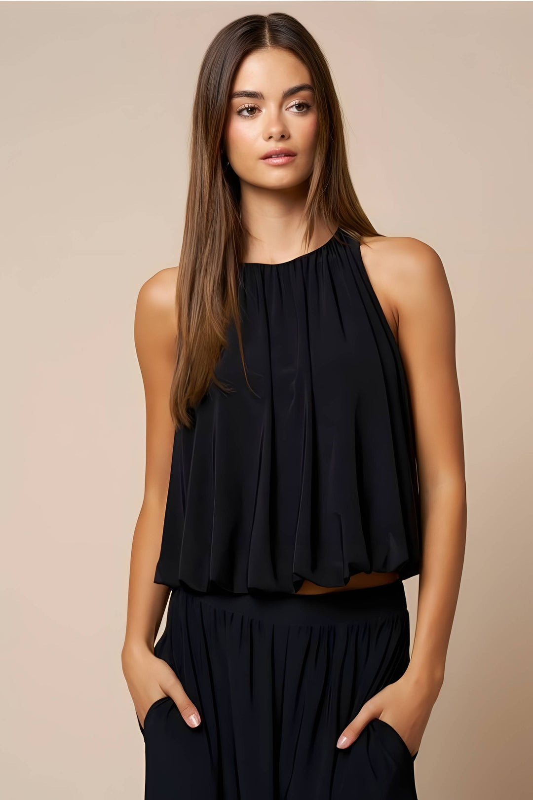 A woman modeling a sleeveless black pleated top with a high gathered neckline, paired with matching black bottoms, against a solid beige studio background.