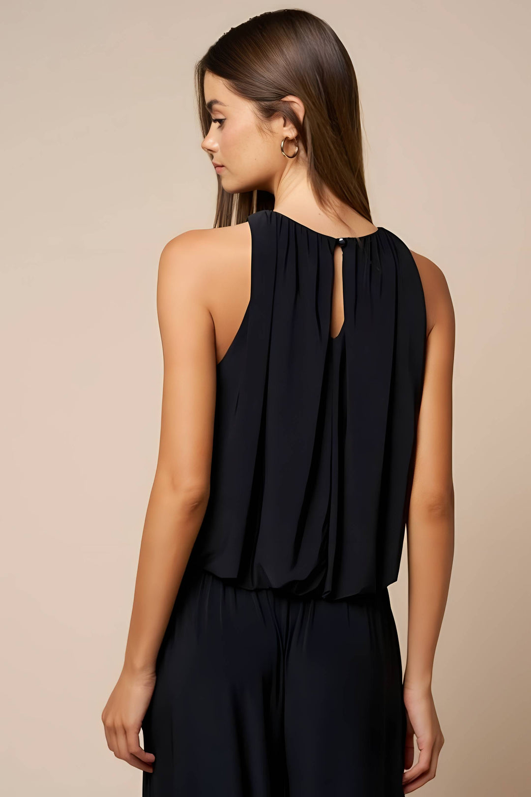 A woman shown from the back wearing a sleeveless black top with a keyhole cutout and pleated back detail, paired with dark bottoms, against a plain beige studio background.