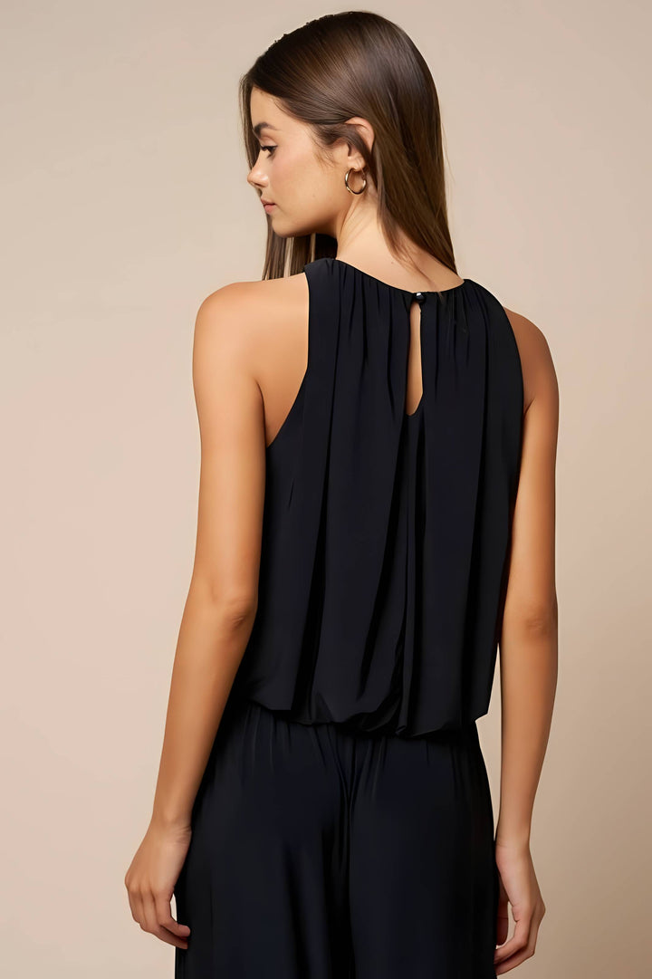 A woman shown from the back wearing a sleeveless black top with a keyhole cutout and pleated back detail, paired with dark bottoms, against a plain beige studio background.