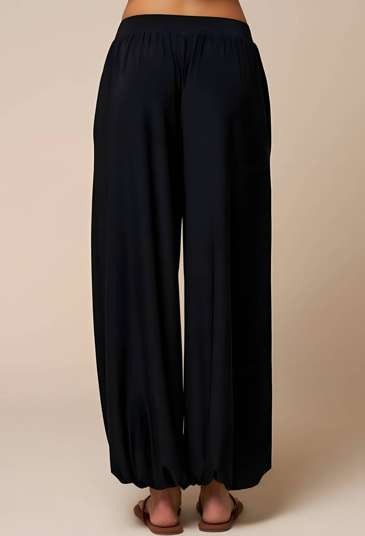 Black wide-leg harem pants shown from the back on a model, featuring an elastic waistband and gathered legs, paired with brown flat sandals against a solid beige background.