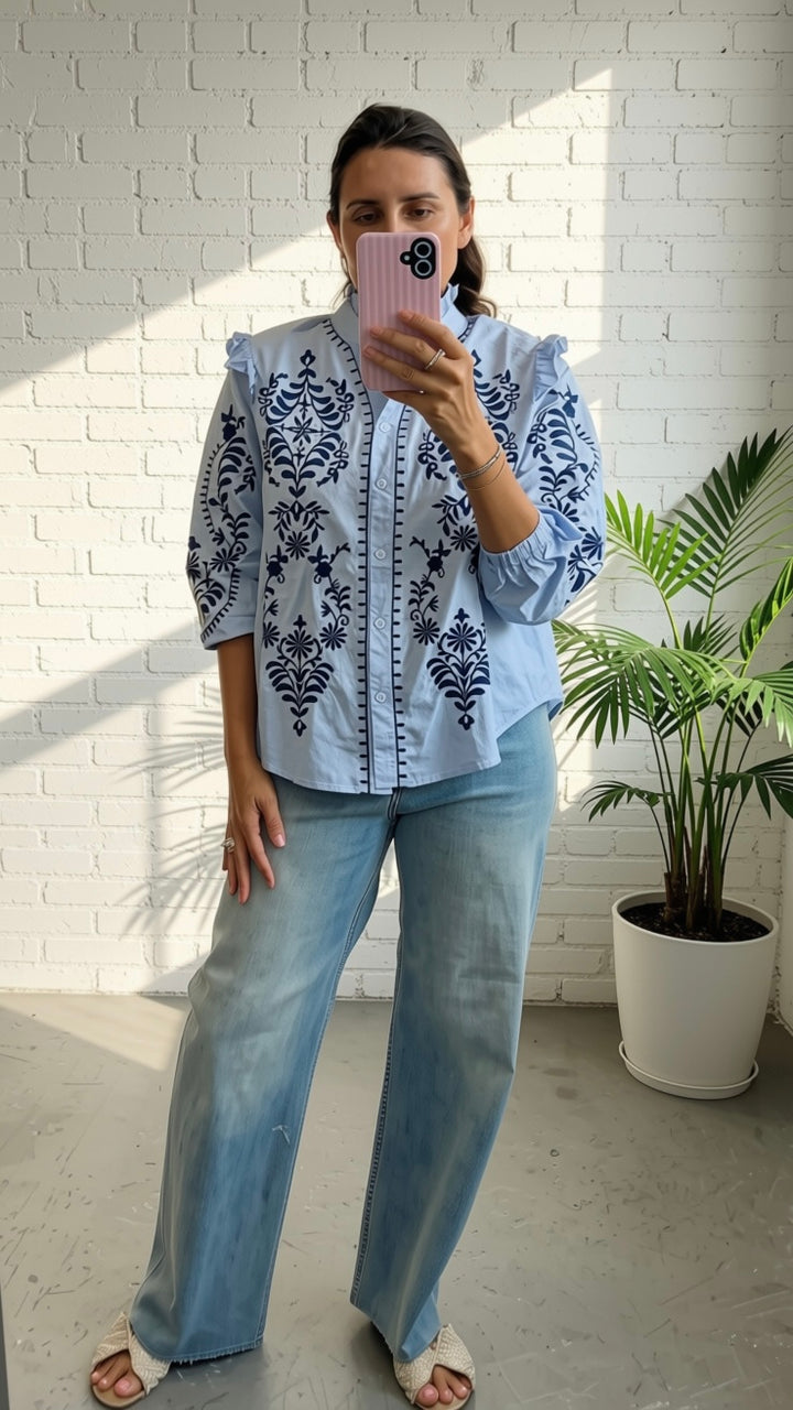 A woman takes a mirror selfie wearing a light blue embroidered blouse with dark blue floral patterns and light-wash wide-leg jeans, standing in front of a white brick wall next to a potted plant.