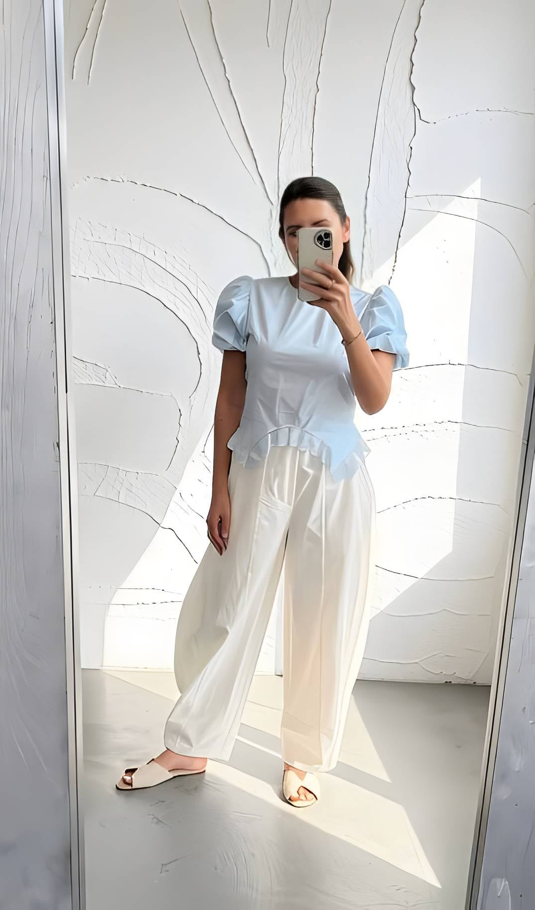 A woman takes a mirror selfie wearing a light blue puff-sleeve top and cream wide-leg trousers, standing in a brightly lit room with a textured white wall.