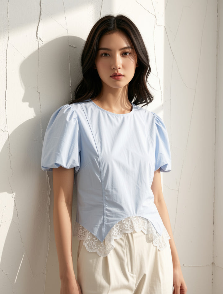 A woman wearing a light blue puff-sleeve blouse with a lace hem detail, paired with cream-colored pants, standing against a white textured wall with soft sunlight and shadows.