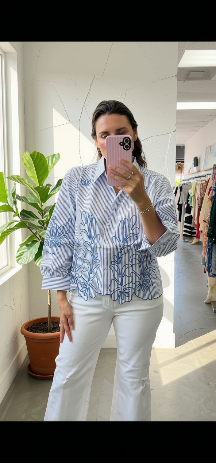 A woman takes a mirror selfie in a boutique, wearing a light blue and white striped button-up shirt with blue floral embroidery on the front, paired with white pants.